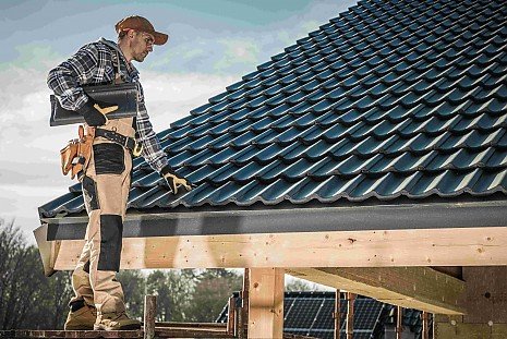 Ein Handwerker in Arbeitskleidung steht auf einem Gerüst an einem neu gedeckten, dunklen Hausdach und prüft die Dachziegel im Zuge einer Dachsanierung. Ein Handwerker in Arbeitskleidung steht auf einem Gerüst an einem neu gedeckten, dunklen Hausdach und prüft die Dachziegel im Zuge einer Dachsanierung.