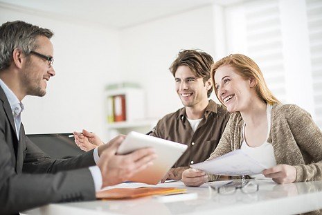 Ein lächelndes junges Paar im Beratungsgespräch mit einem Experten in einem hellen Büro; im Vordergrund wird ein Tablet zur Veranschaulichung genutzt Ein lächelndes junges Paar im Beratungsgespräch mit einem Experten in einem hellen Büro; im Vordergrund wird ein Tablet zur Veranschaulichung genutzt